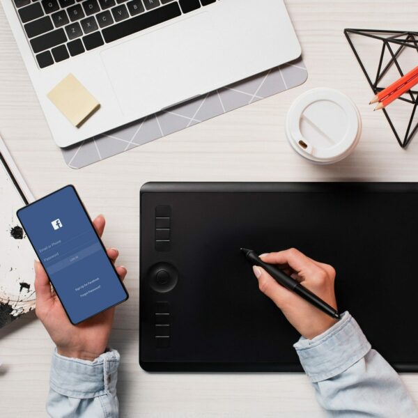 cropped view of designer using graphics tablet, pen and smartphone with facebook on screen, flat lay